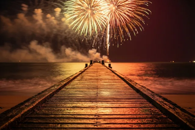 Een houten pier loopt de zee in onder kleurrijk vuurwerk, met warm oranje en geel licht dat weerspiegelt op het water.