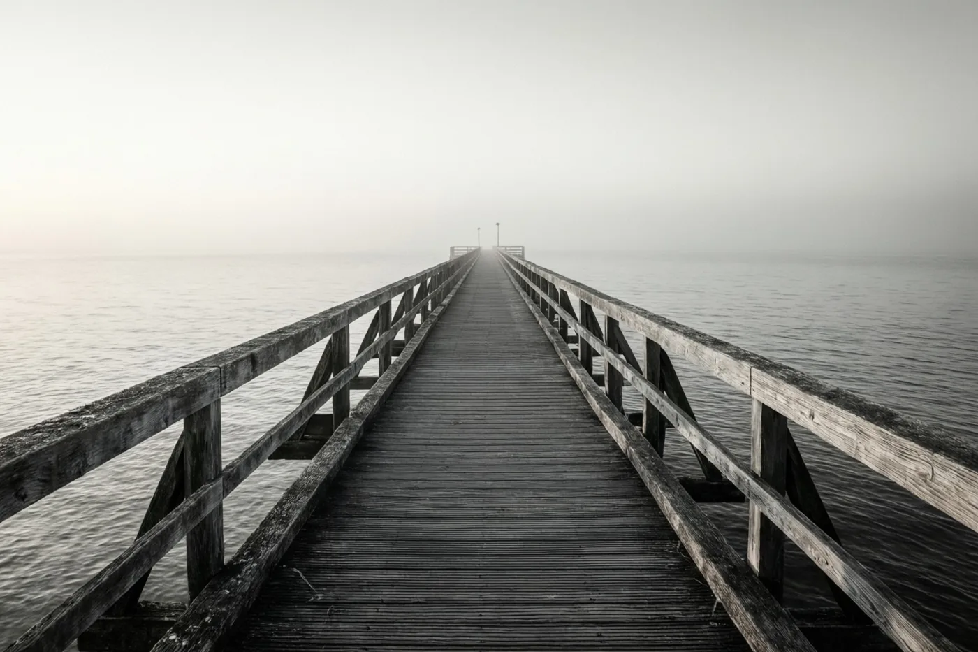 Een lange houten pier strekt zich uit in een kalme zee onder een nevelige hemel, in gedempte tinten.