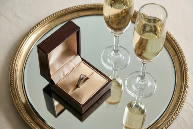 Een open houten ringdoosje met een diamanten verlovingsring staat naast twee champagnefluiten op een spiegelend dienblad met gouden rand, op een lichtgekleurd tafelkleed.