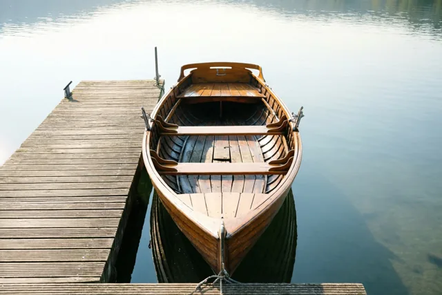 Een oude houten roeiboot ligt aan een houten steiger op een stil meer in gedempte bruine, grijze en blauwe tinten.