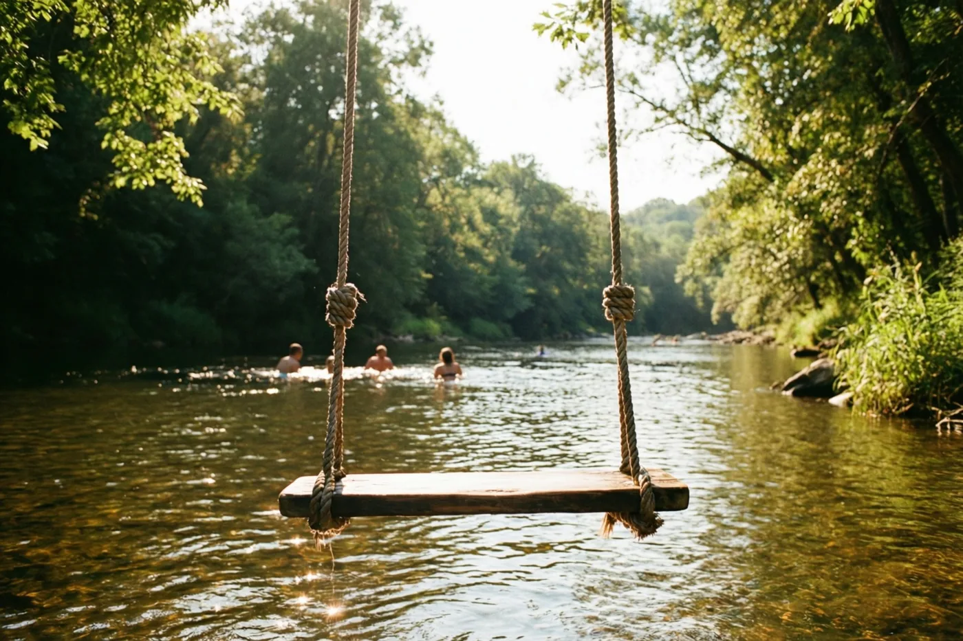 Een houten schommel aan touwen hangt boven een zonnige rivier met meerdere zwemmers op de achtergrond en weelderige groene bomen langs het water.