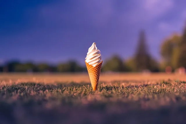 Een hoorntje met softijs staat rechtop in het gras onder een paars-oranje schemerlucht, in warm goudkleurig licht.