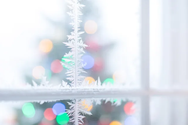 IJskristallen vormen fijne patronen op een ruit, met wazige gekleurde kerstlichtjes op de achtergrond in witte en zachte pasteltinten.