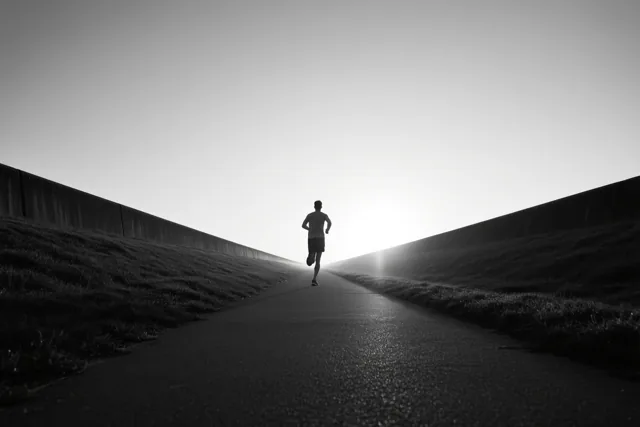 Een eenzame jogger loopt weg over een verhard dijkpad bij zonsopgang, in zwart-wit met een donkere weg en een heldere hemel.