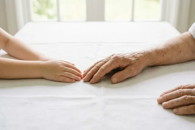 Een heel jonge hand en een oudere hand rusten samen op een wit linnen tafelkleed, met zacht daglicht uit een raam op de achtergrond.