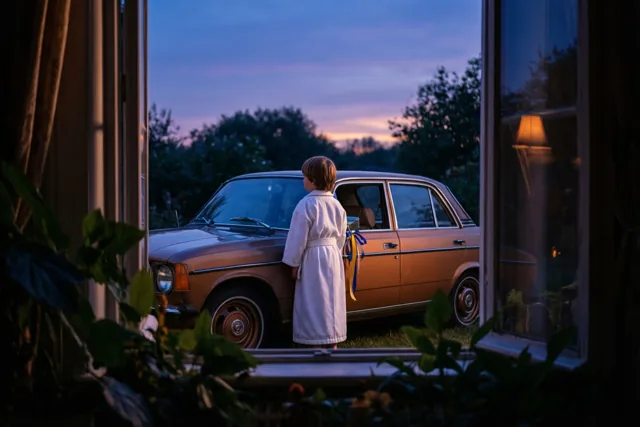 Door een open raam is een jonge jongen in een wit gewaad te zien naast een bruine auto uit de jaren zeventig met linten, onder een paarse en oranje schemerlucht.