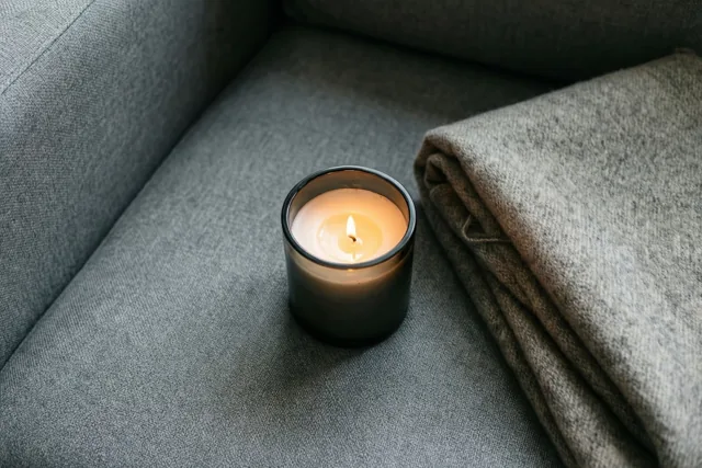 Een brandende kaars in een houder van rookglas staat op een grijze stoffen zetel naast een opgevouwen wollen deken.