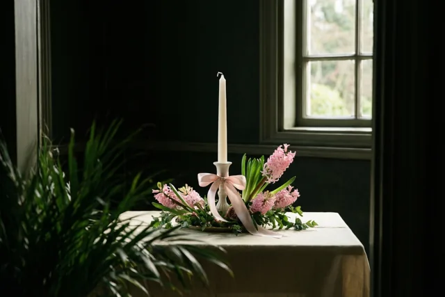 Een witte kaars met een roze lint staat tussen roze hyacinten op een tafel met een licht kleed, bij een raam in een kamer met donkere muren.