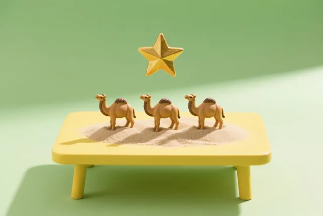 Drie kameelbeeldjes staan op zand op een gele tafel, met een gouden ster erboven tegen een groene achtergrond.