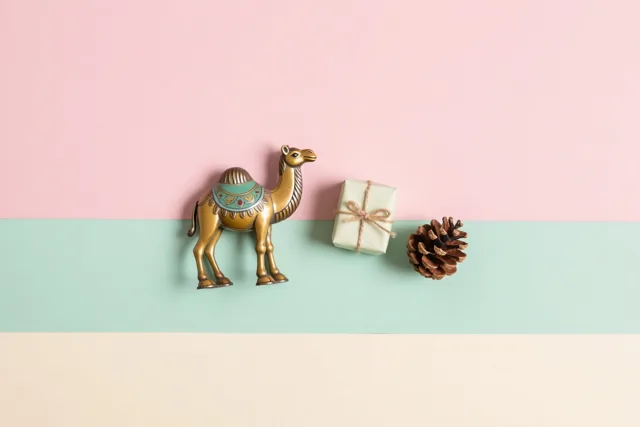 Een antiek blikken kameeltje staat naast een klein ingepakt cadeau en een dennenappel voor horizontale roze, mintgroene en crèmekleurige banen.
