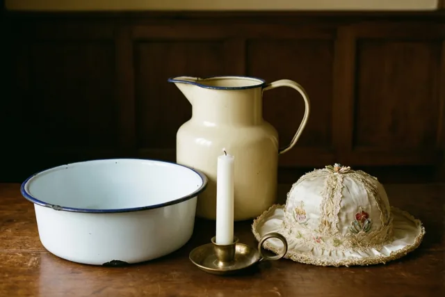 Een oude emaille kan en kom staan op een houten tafel naast een witte doopkaars in een messing kandelaar en een geborduurd doopmutsje, voor een donkere houten wand.