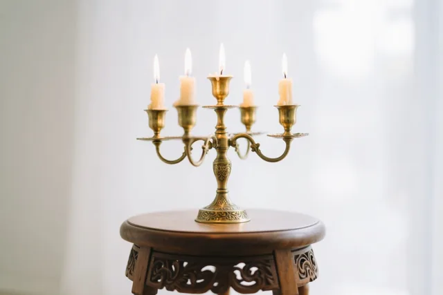 Een messing kandelaar met vijf brandende kaarsen staat op een houten krukje met snijwerk tegen een heldere, onscherpe achtergrond.