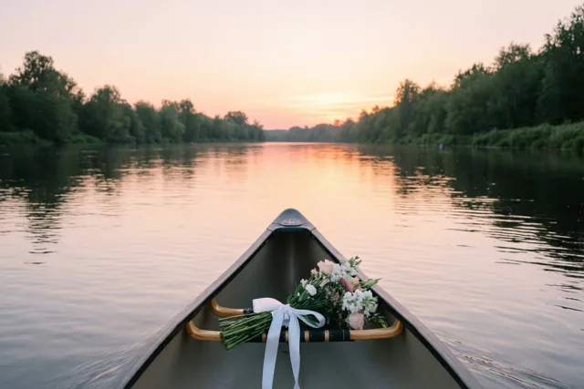Een kano met een boeket witte en roze bloemen en een wit lint ligt vooraan op een rustige rivier tussen bomen onder een roze en oranje avondlucht.