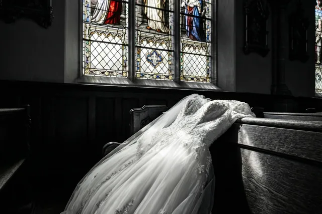 Een wit doopkleed met kanten details ligt over een donkere houten kerkbank, verlicht door een glasraam in een schemerige kerk met houten lambrisering en religieuze kunst.