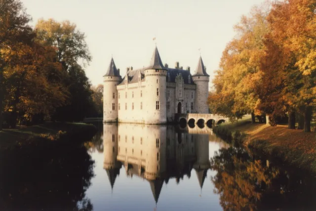Een Vlaams kasteel met slotgracht weerspiegelt in rustig water tussen bomen met gouden herfstbladeren in zacht licht.