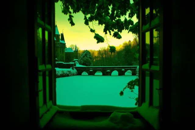 Door een open raam met groenige bladeren eromheen is een besneeuwde kasteelgracht te zien met een oude stenen brug in de verte, in groen en geel licht.