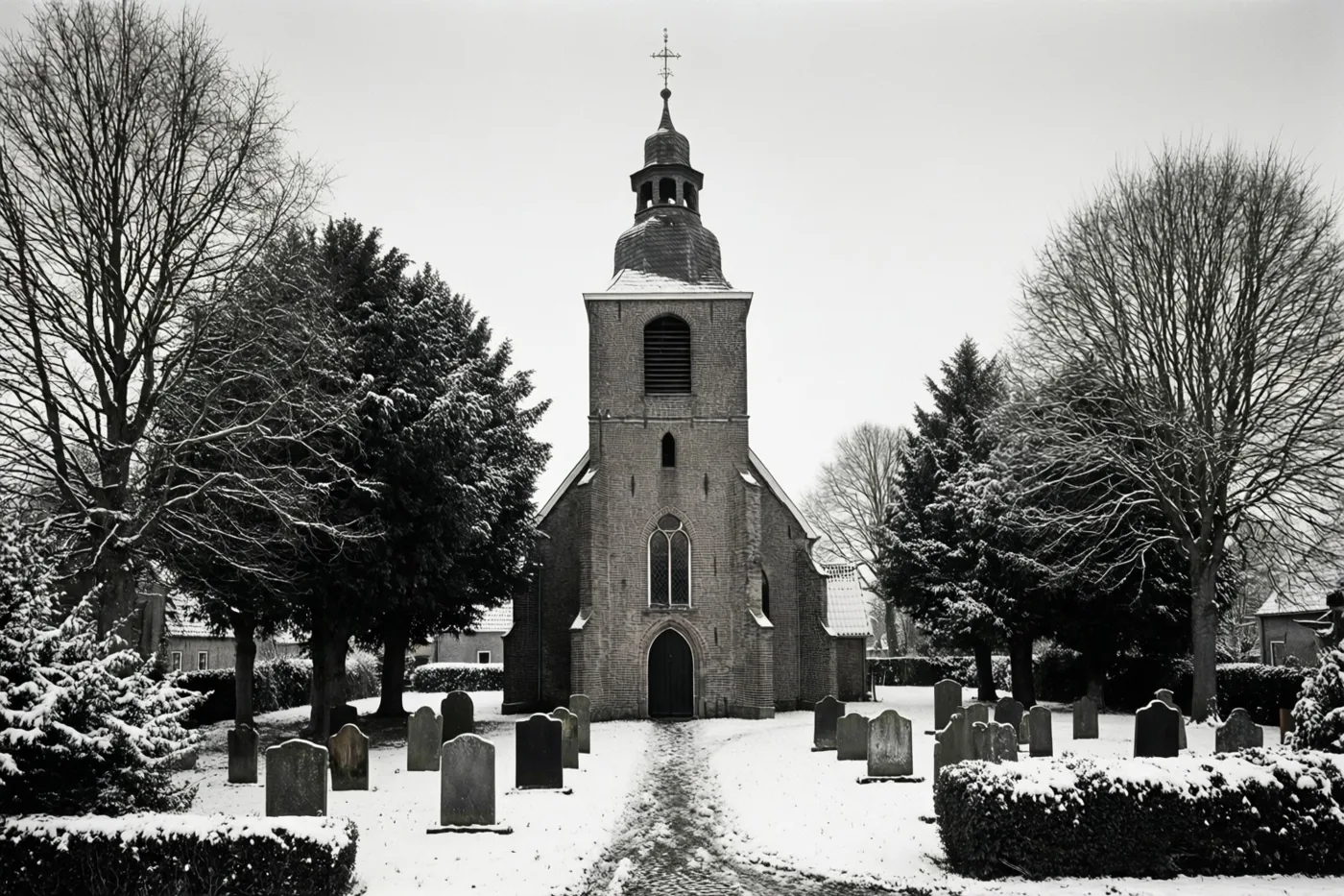 Een kleine bakstenen kerk met een toren en een kruis staat op een besneeuwd kerkhof tussen besneeuwde bomen en grafstenen, weergegeven in zwart-wit.