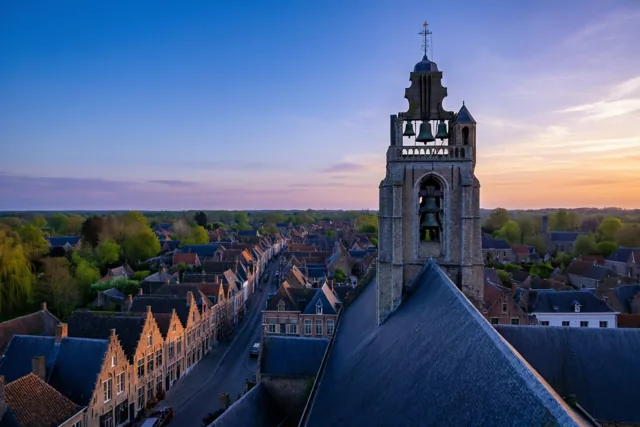 Een hoge stenen kerktoren met zichtbare klokken kijkt uit over een straat met traditionele huizen in een Vlaams dorp bij schemering, onder een blauwe lucht met oranje aan de horizon.