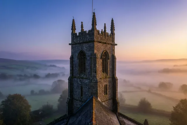 Een stenen kerktoren steekt bij dageraad boven de mist in een landelijk landschap uit, onder een hemel die van zachtblauw naar oranje verloopt.