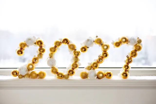 Gouden en witte kerstballen vormen 2027 op een besneeuwde vensterbank tegen een heldere witte achtergrond.