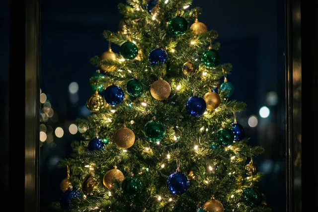 Door een donker raam is een kerstboom met groene, blauwe en gouden versiering en warme lichtjes te zien, met onscherpe lichtvlekken op de achtergrond.