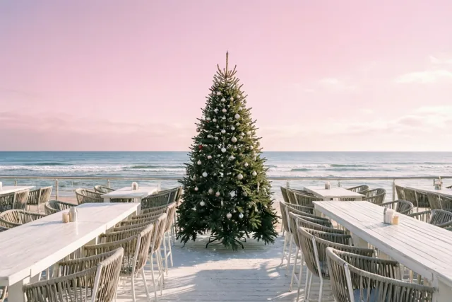 Een versierde kerstboom staat op het houten terras van een strandbar tussen witte tafels en stoelen, met de zee en een roze lucht op de achtergrond.