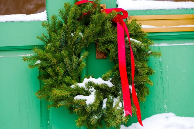 Een groene kerstkrans met een rood lint hangt aan een verweerde groene houten deur met een dun laagje sneeuw.
