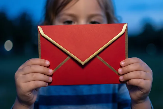 Een klein kind houdt een rode envelop met een gouden rand voor het gezicht, tegen een wazige blauwe avondachtergrond.