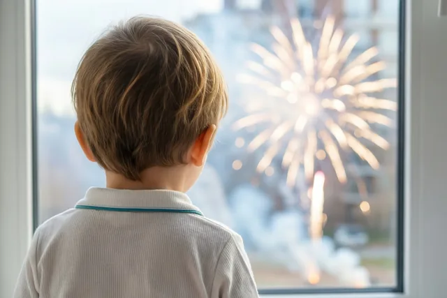 Een jong kind in een lichtgekleurde pyjama kijkt met de rug naar de camera door een raam naar helder, onscherp vuurwerk.
