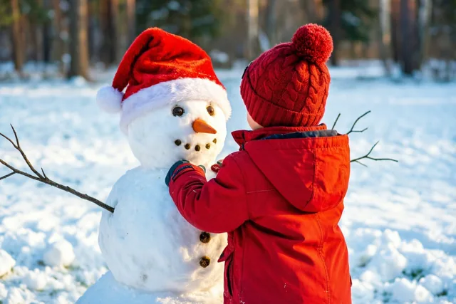 Een kind in een rode jas en pomponmuts zet van achteren gezien een tak op een sneeuwman met kerstmuts in een zonnig, besneeuwd bos.