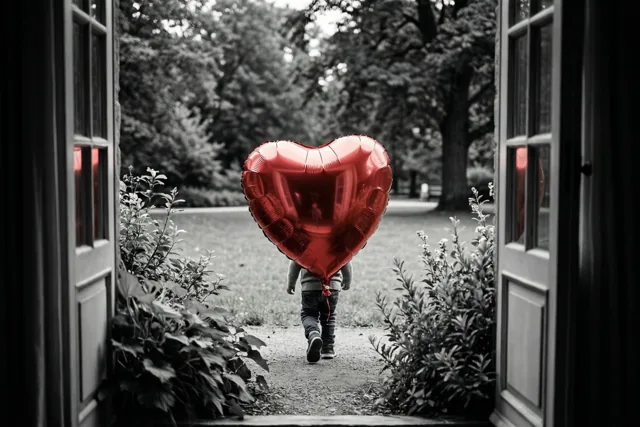 Door een open deuropening is een kind van achteren te zien dat met een grote rode hartvormige ballon over een parkpad wegloopt, tegen een zwart-witte achtergrond.