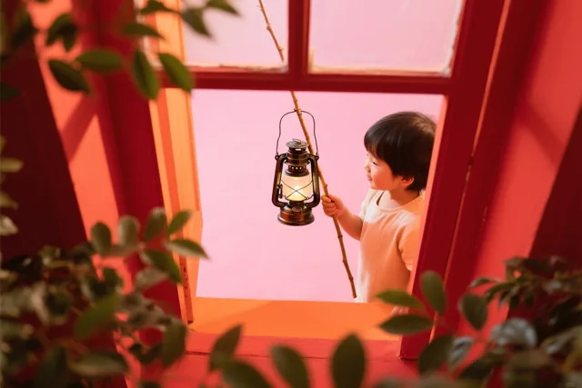 Een jong kind staat in een rood raamkozijn en houdt een brandende lantaarn aan een stok vast, in warm rood en roze licht.