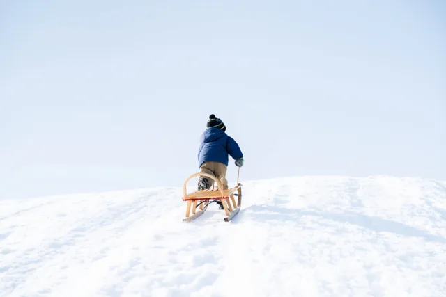 Een kind in een blauwe jas en gestreepte muts trekt, van achteren gezien, een houten slee bergop door diepe sneeuw onder een heldere, lichtblauwe lucht.