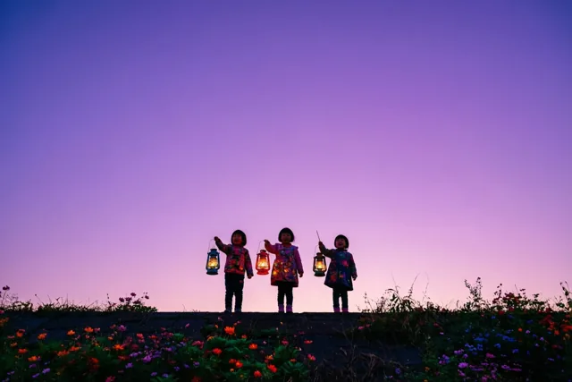 Drie kleine kinderen staan als silhouetten met elk een brandende lantaarn op een dijk tegen een paarse schemerhemel, met kleurrijke veldbloemen op de voorgrond.