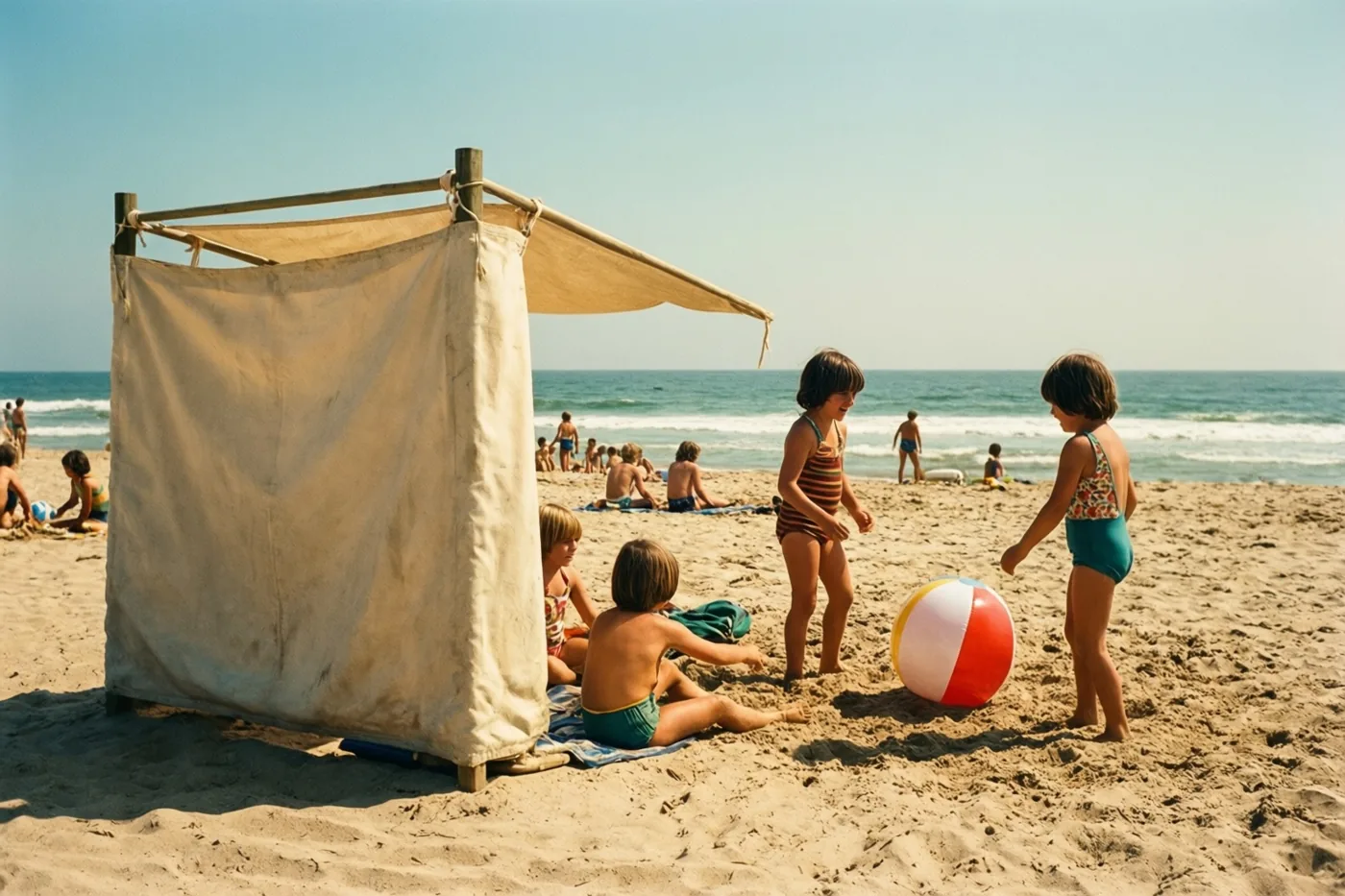 Kinderen in zwemkleding uit de jaren zeventig spelen met een gestreepte strandbal naast een canvas windscherm op een zonnig strand, met de zee onder een helderblauwe hemel.
