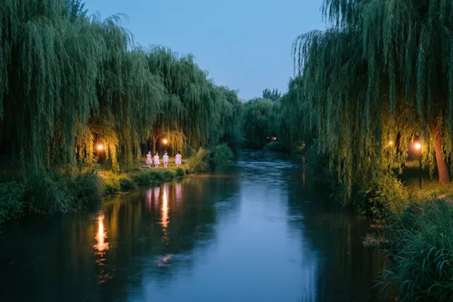 Vier kinderen in lichtgekleurde jurken staan met lantaarns aan een rivieroever onder groene wilgen, met warme weerspiegelingen op het water en een diepblauwe schemerhemel.
