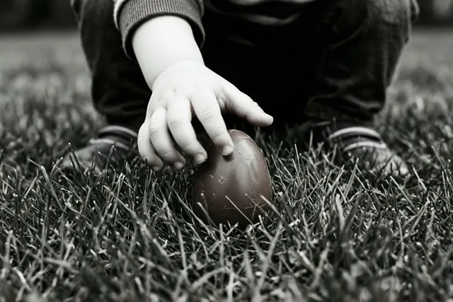 Een kinderhand reikt naar een chocolade-ei in het gras op een zwart-witfoto.