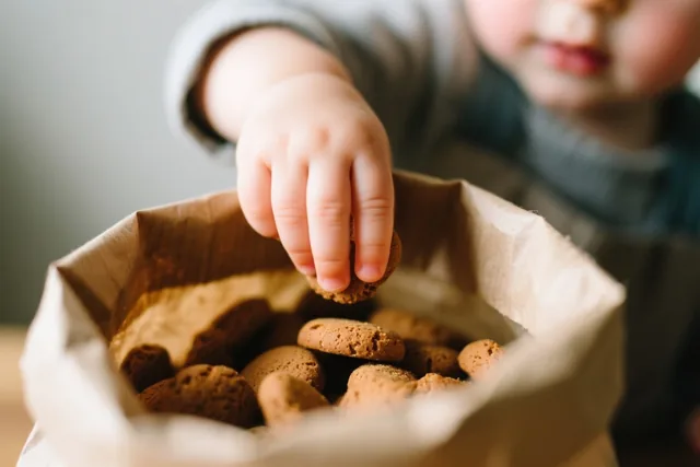 Een kinderhand reikt in een papieren zak vol bruine pepernoten, in zacht warm licht.