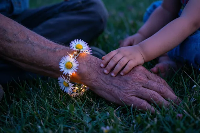 Een kinderhand ligt op de gerimpelde pols van een oudere persoon met een madeliefjesarmband, op gras in koel, gedempt avondlicht.