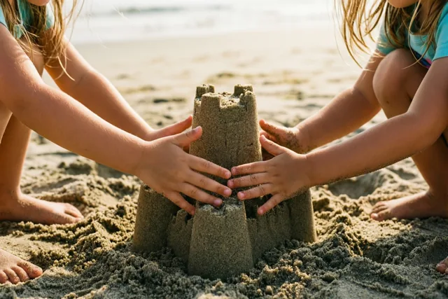 De handen van twee kleine kinderen drukken samen een zandkasteel aan op een strand in warm goudkleurig zonlicht.