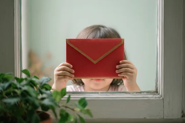 Door een raamkozijn zijn kinderhandjes te zien die een rode envelop met een gouden rand vasthouden, met een groene plant op de voorgrond.