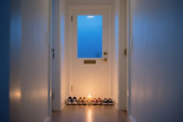 In een gang staan kinderschoenen netjes naast een witte voordeur met blauwgetint glas, verlicht door één brandende kaars op de vloer.