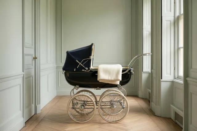 Een klassieke kinderwagen met donkerblauwe kap en ivoorkleurig wollen deken staat in een hal met lichtgroene muren en een houten visgraatvloer, verlicht door een raam.