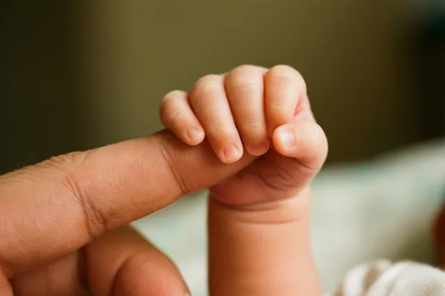 Een pasgeboren baby grijpt met een klein handje een vinger van een volwassene vast tegen een onscherpe achtergrond in warme, gedempte tinten.