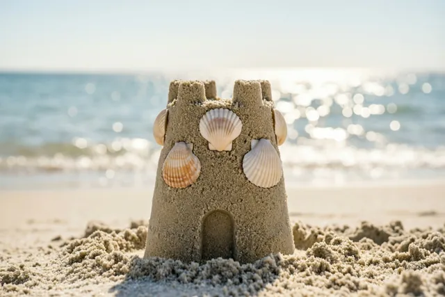 Een klein zandkasteel versierd met schelpen staat op een zandstrand, met een glinsterende zee en heldere hemel erachter in warm zonlicht.