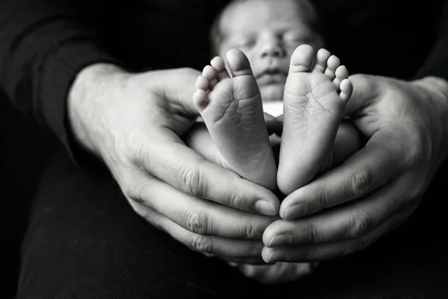 Een zwart-witfoto toont de voetjes van een pasgeboren baby in de handen van een volwassene tegen een donkere achtergrond.