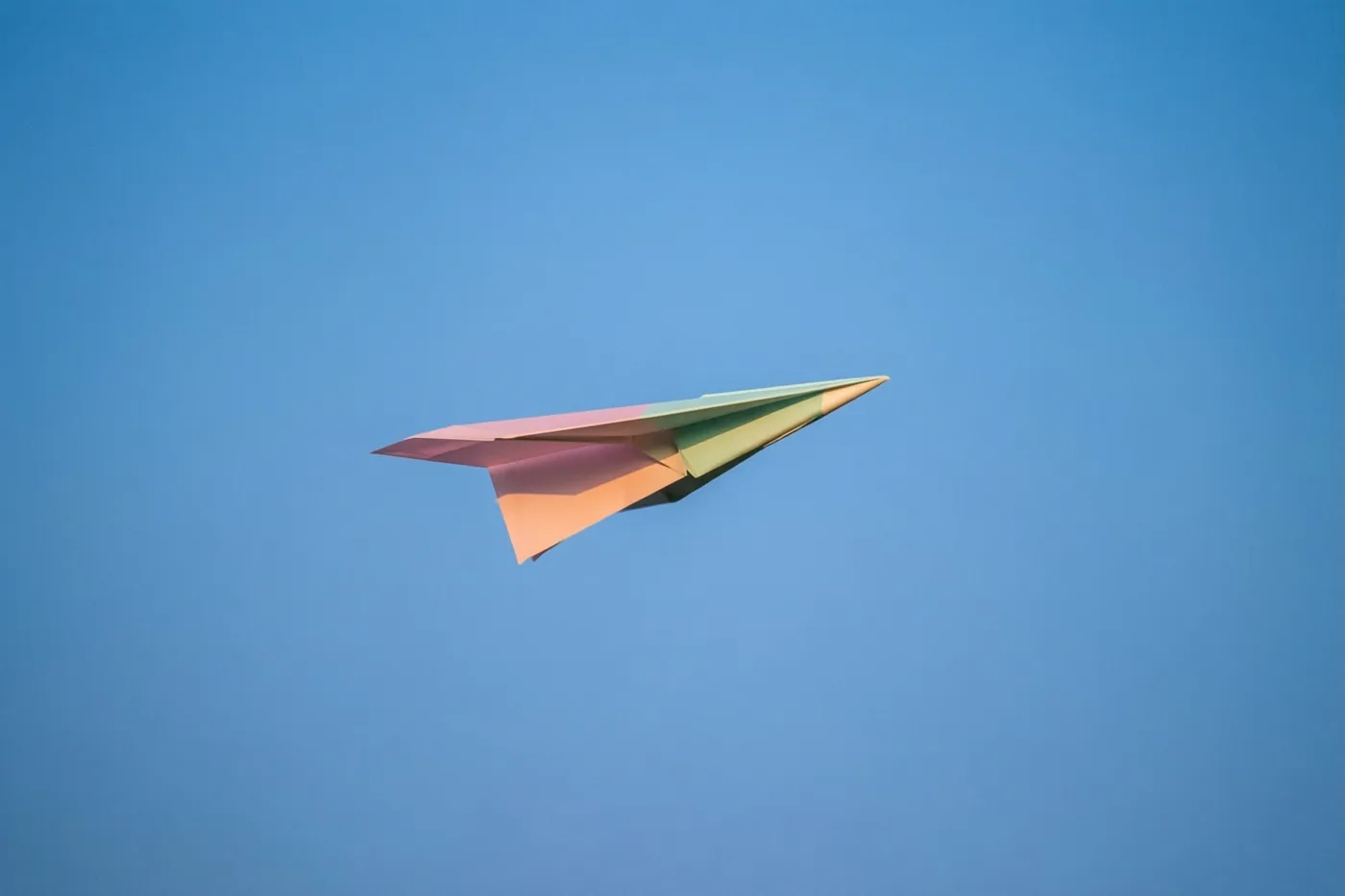 Een kleurrijk papieren vliegtuigje met blauw, roze en groen vliegt door een heldere blauwe lucht.
