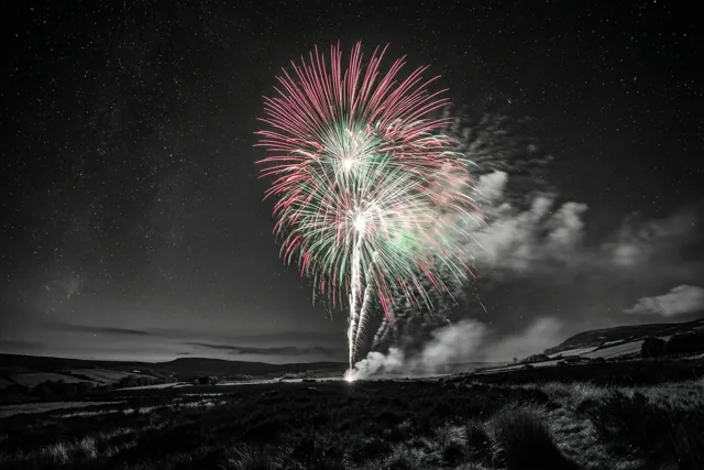 Kleurrijk vuurwerk barst los boven een donkere heide met sterren en vage wolken, terwijl de rest van het beeld in zwart-wit is weergegeven.