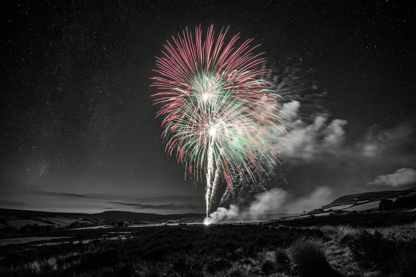 Kleurrijk vuurwerk barst los boven een donkere heide met sterren en vage wolken, terwijl de rest van het beeld in zwart-wit is weergegeven.