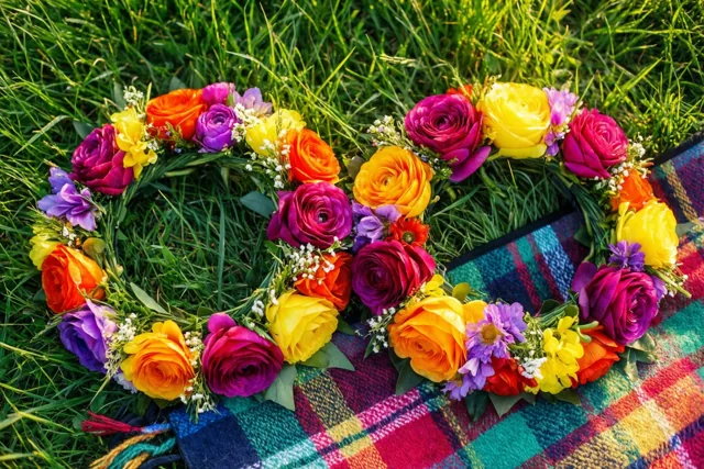 Twee kleurrijke bloemenkronen met gele, magenta en oranje tinten liggen naast elkaar op een geruit picknickdeken op groen gras.